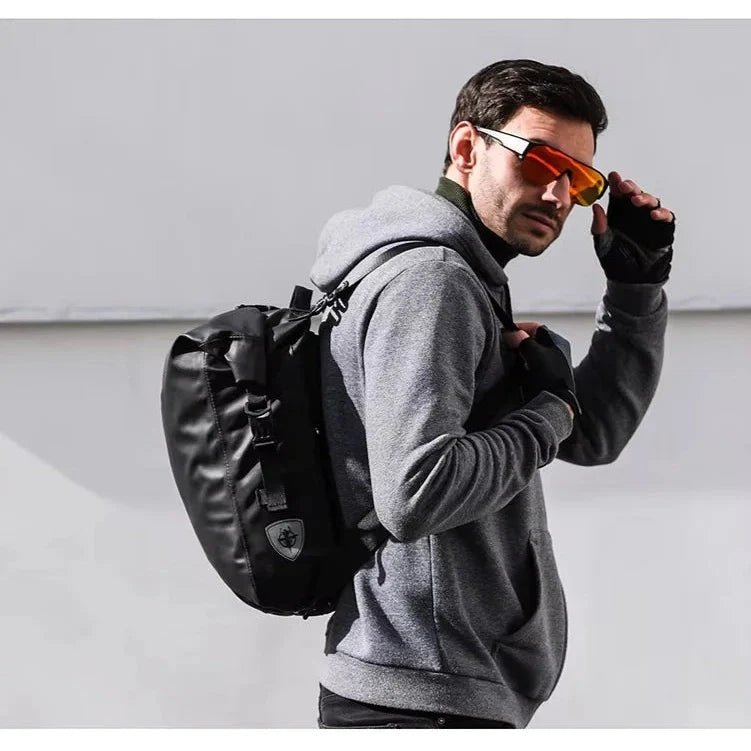 Homme avec hoodie gris, gants noirs, sac à dos imperméable vélo, tenant des lunettes de soleil rouge